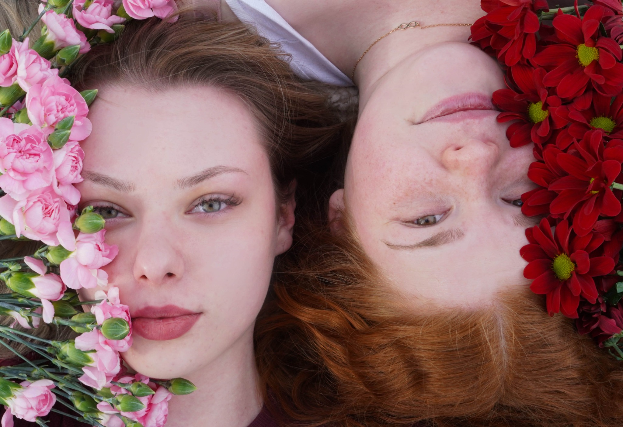 Portret dwóch młodych kobiet, leżących obok siebie, z twarzami otoczonymi kwiatami: różowe goździki i czerwone chryzantemy, delikatny makijaż, ciepłe oświetlenie.