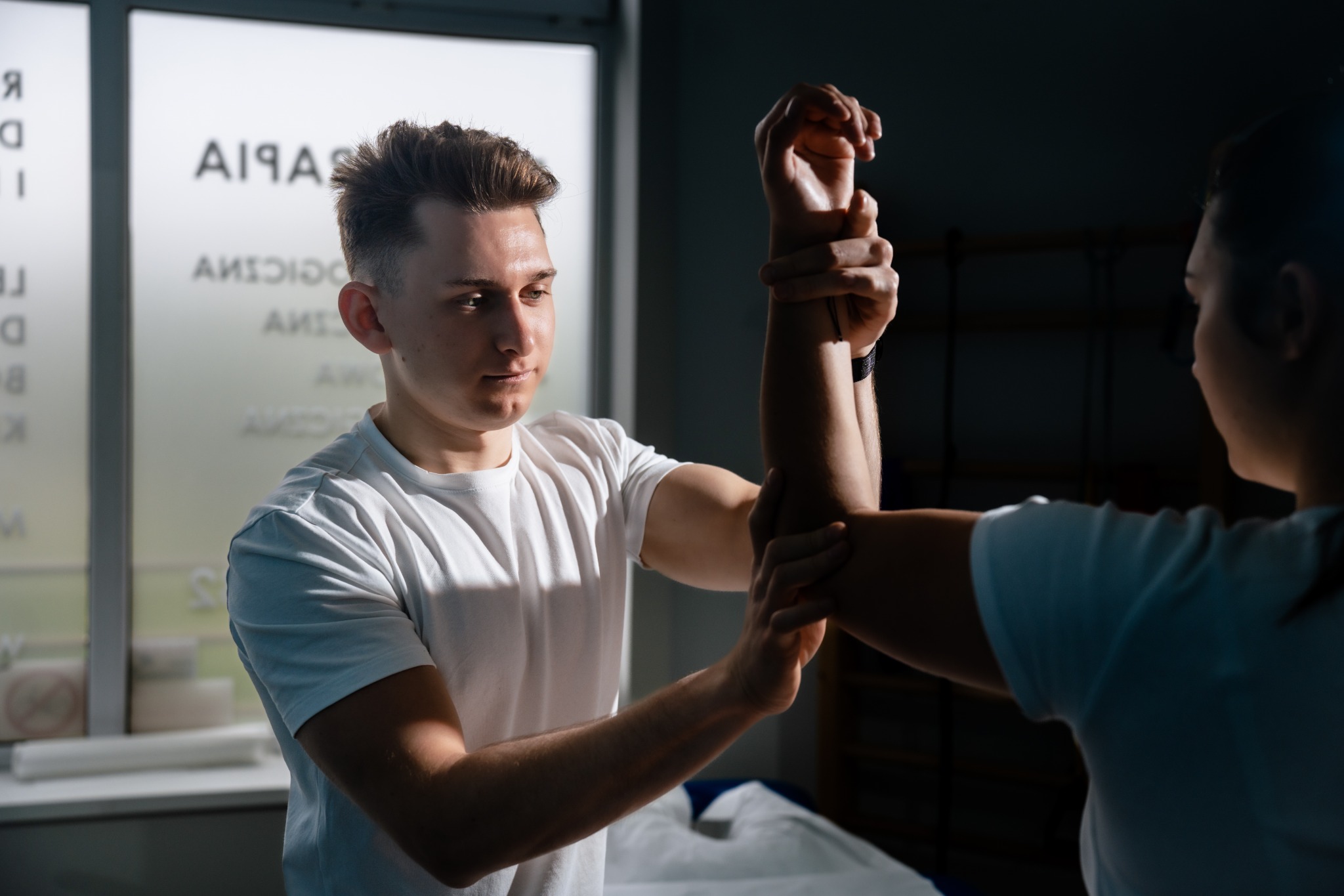 Mężczyzna w białej koszulce bada zakres ruchu ramienia pacjentki w gabinecie, w tle okno z napisami.