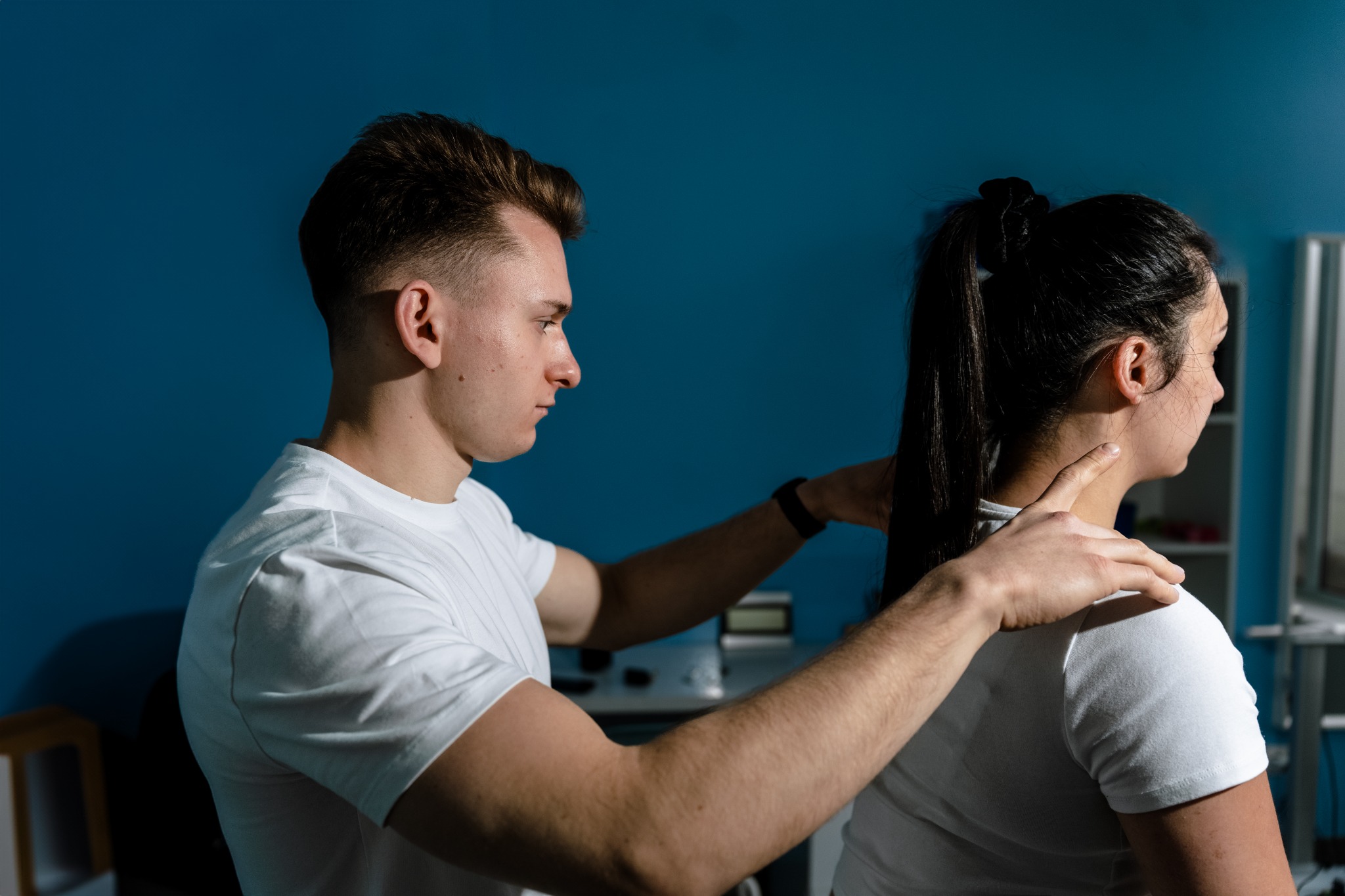 Mężczyzna w białej koszulce wykonuje badanie palpacyjne szyi kobiety w gabinecie o błękitnych ścianach.