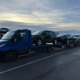 Niebieska laweta przewozi cztery samochody osobowe, w tym czarny Range Rover, na tle pochmurnego nieba o świcie.