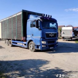 O-VIA TRANSPORT SP. Z O.O. - Niebieski samochód ciężarowy marki MAN z naczepą na żwirowym placu, widoczne cienie drzew i inne pojazdy w tle.