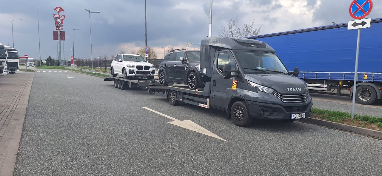 Czarna laweta Iveco z dwoma samochodami (białe BMW i czarne Mini Cooper) na parkingu przy drodze, w tle widoczny szyld Orlen i KFC.