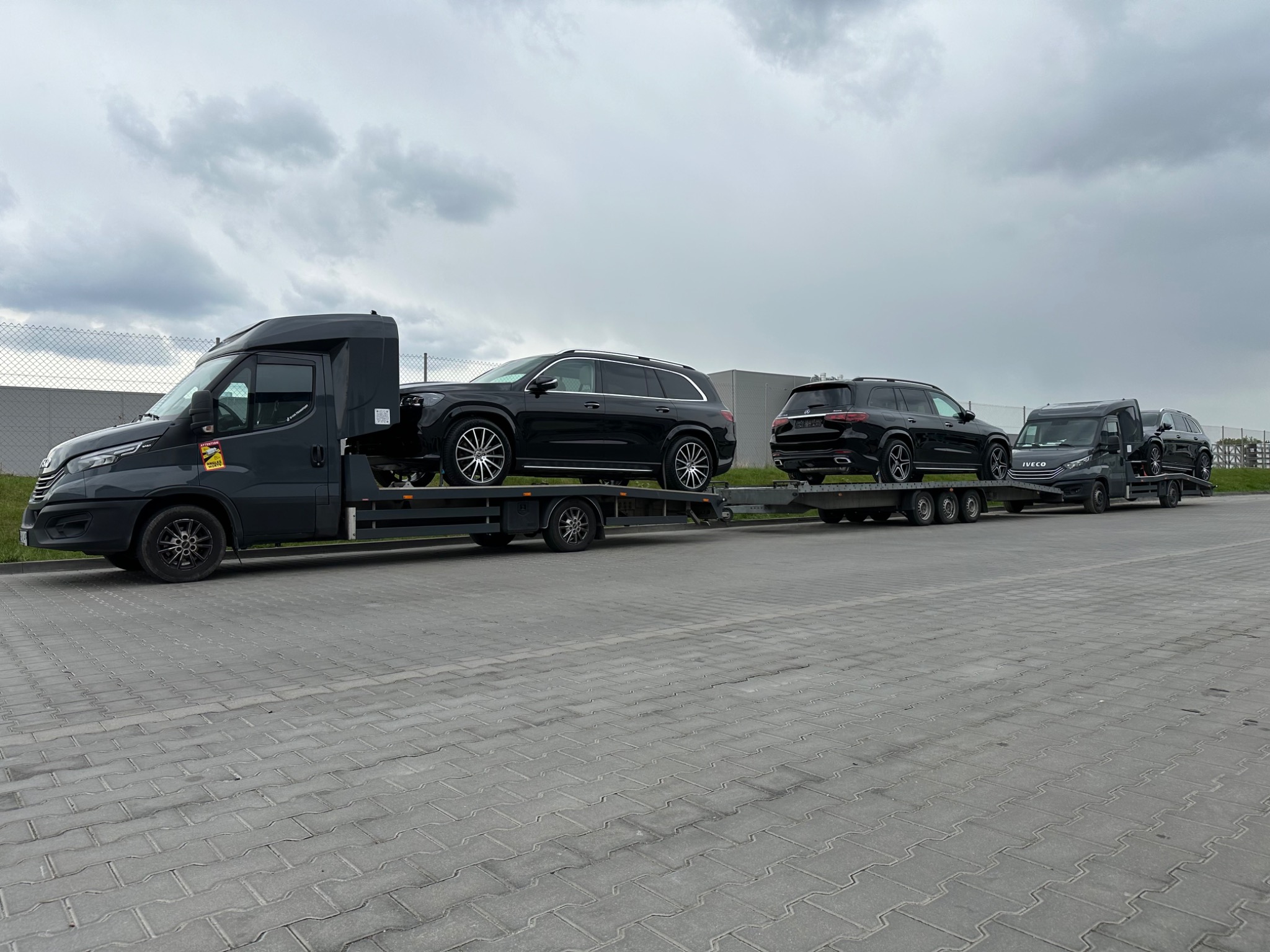 Długi zestaw lawet z czarnymi samochodami osobowymi typu SUV na platformach, stojący na szarym brukowanym placu pod pochmurnym niebem.