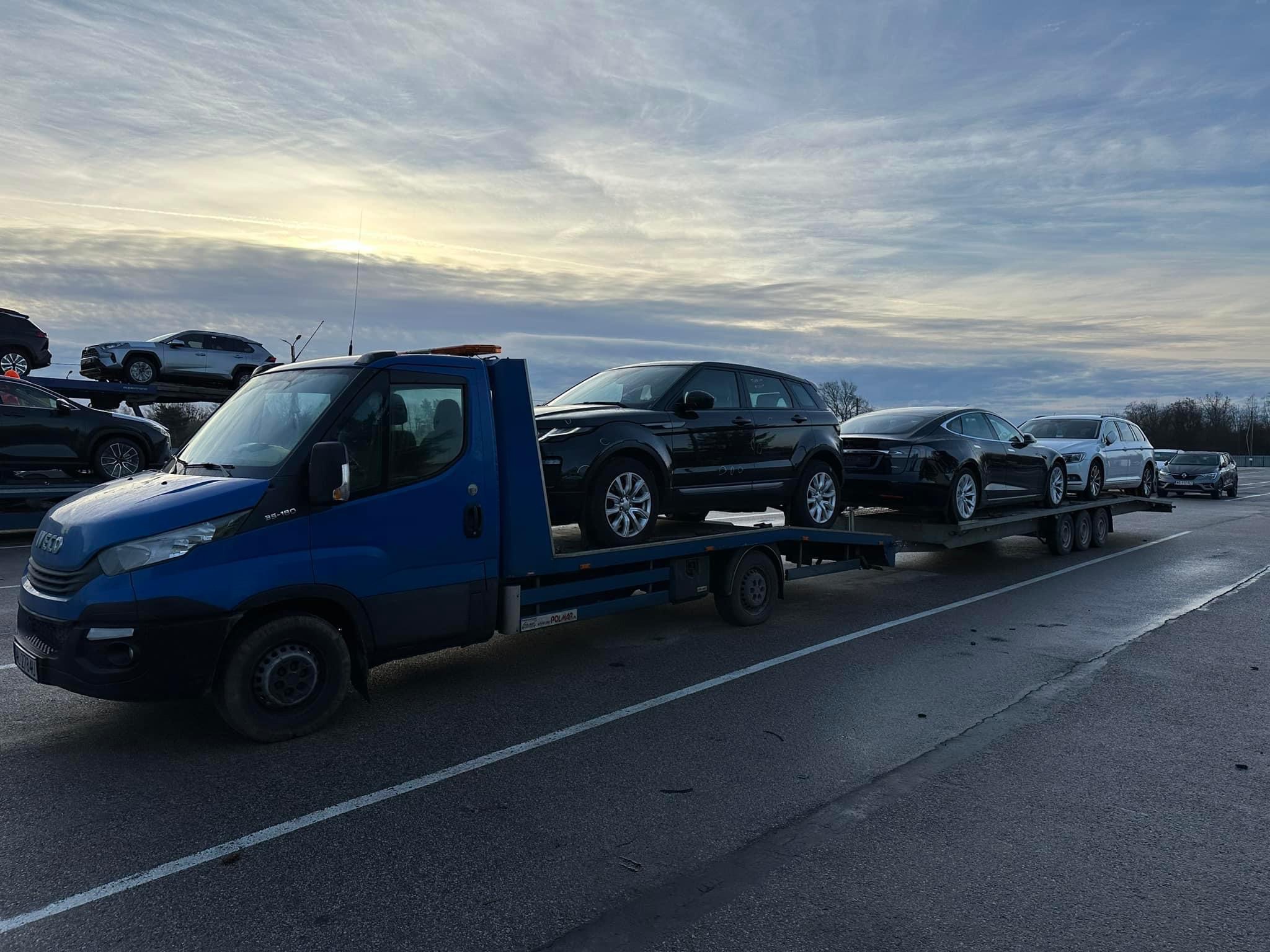 Niebieska laweta przewozi cztery samochody osobowe, w tym czarny Range Rover, na tle pochmurnego nieba o świcie.