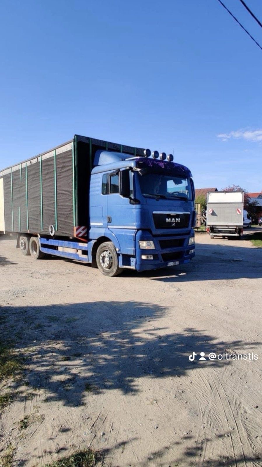 Niebieski samochód ciężarowy marki MAN z naczepą na żwirowym placu, widoczne cienie drzew i inne pojazdy w tle.