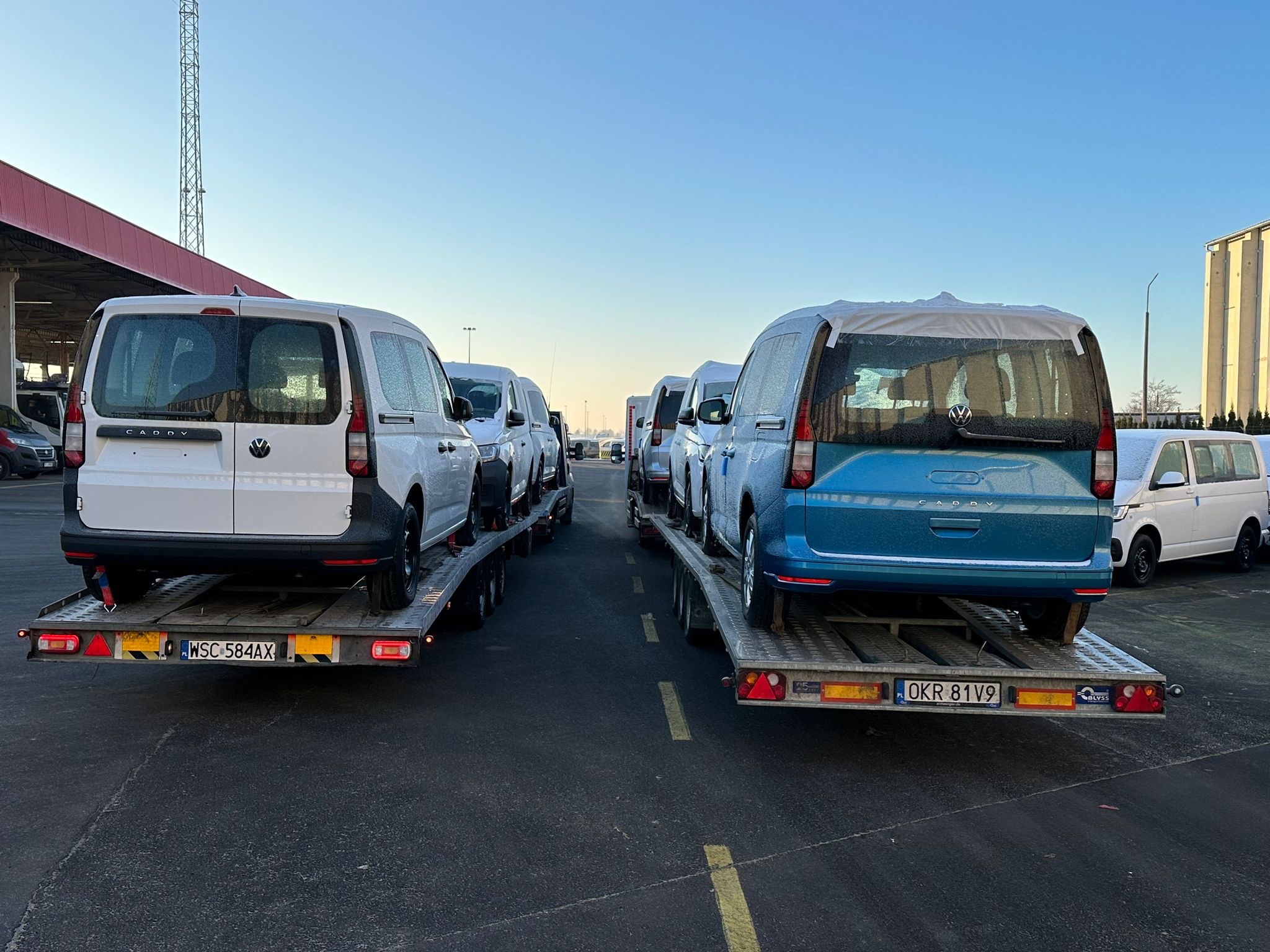 Nowe samochody typu furgon marki Volkswagen Caddy zabezpieczone folią transportowane na lawetach z widocznymi tablicami rejestracyjnymi, na tle placu z budynkami przemysłowymi i wieżą...