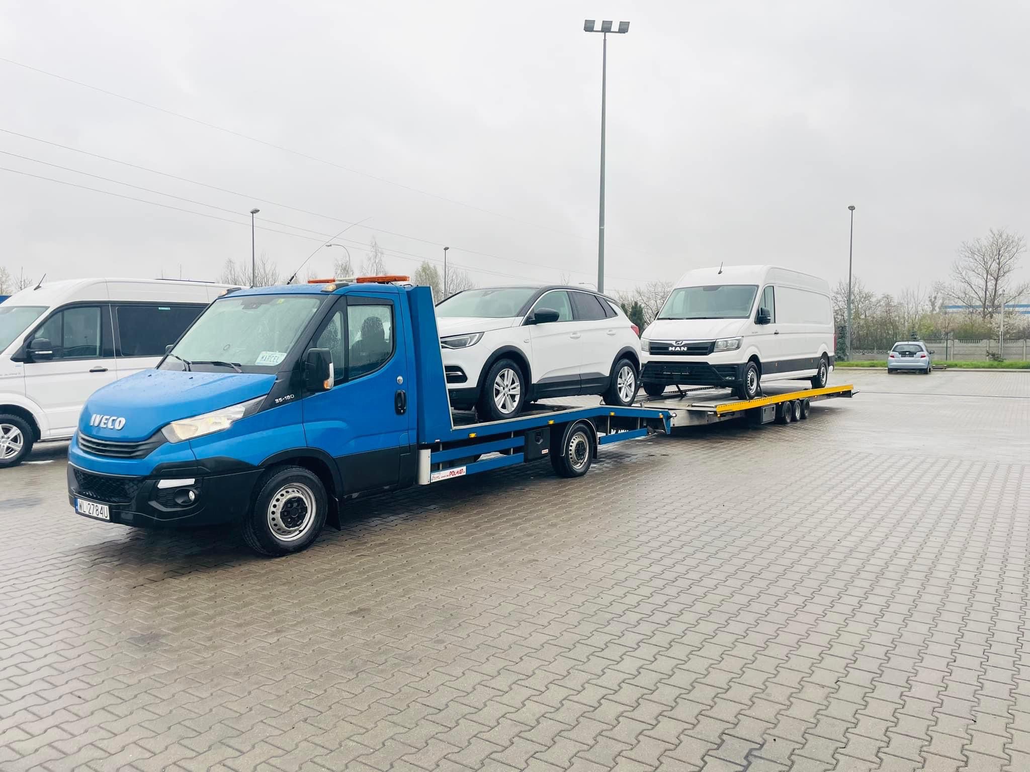 Niebieska laweta Iveco z pomarańczowym światłem ostrzegawczym przewozi białego SUV-a Opel Grandland X i białego vana MAN na parkingu z kostki brukowej w pochmurny dzień.