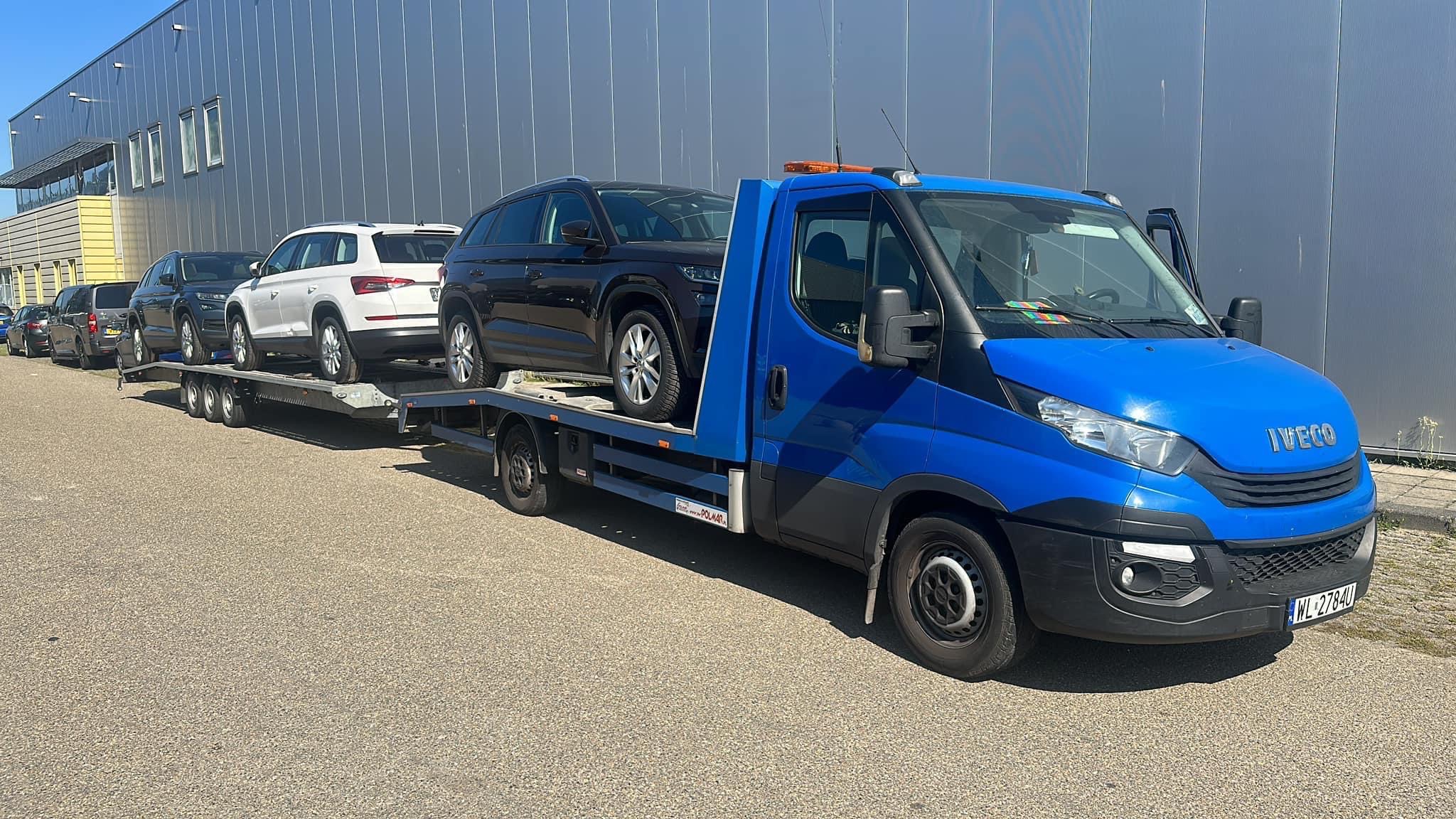 Niebieska laweta Iveco z pomarańczowym światłem ostrzegawczym przewozi trzy samochody osobowe marki Skoda na platformie, zaparkowana na szarym asfalcie przed budynkiem z szarej blachy falistej.