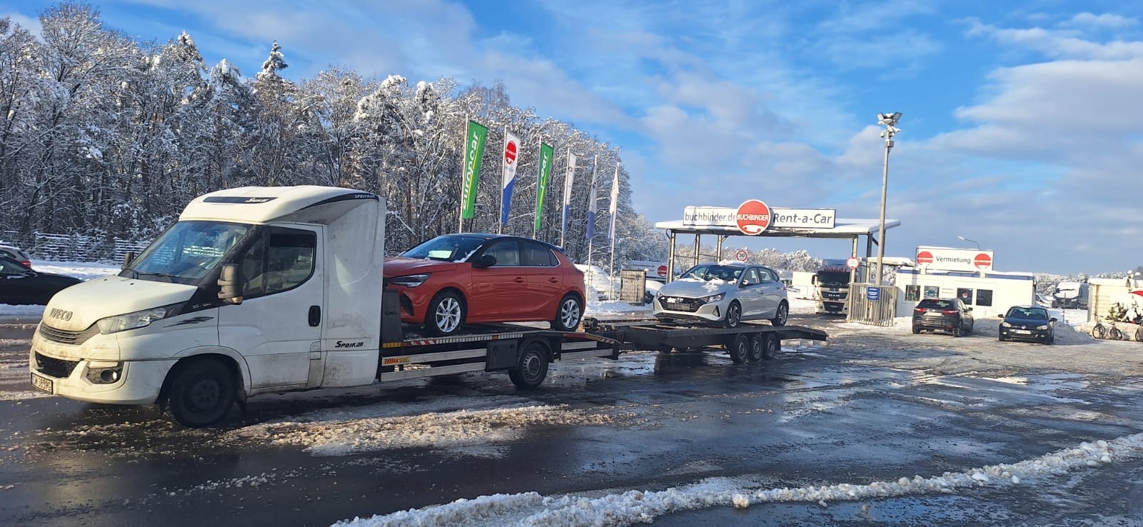 Biała laweta Iveco z pomarańczowym Oplem Corsą na platformie, na tle zasypanego śniegiem parkingu wypożyczalni samochodów Rent-a-Car w zimowy dzień.