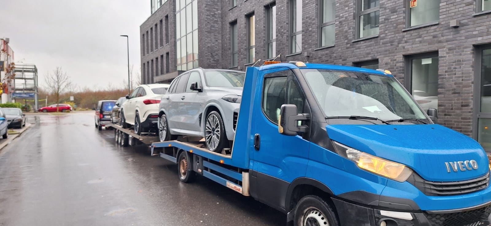 Niebieska laweta Iveco przewozi dwa luksusowe samochody osobowe: biały Mercedes GLE Coupe i srebrne BMW X5 na tle nowoczesnego budynku z cegły.
