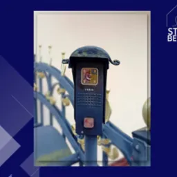 Czarny panel wejściowy z kamerą i przyciskiem dzwonka, model Vidos, zamontowany na metalowym słupku bramy z ozdobnymi elementami.