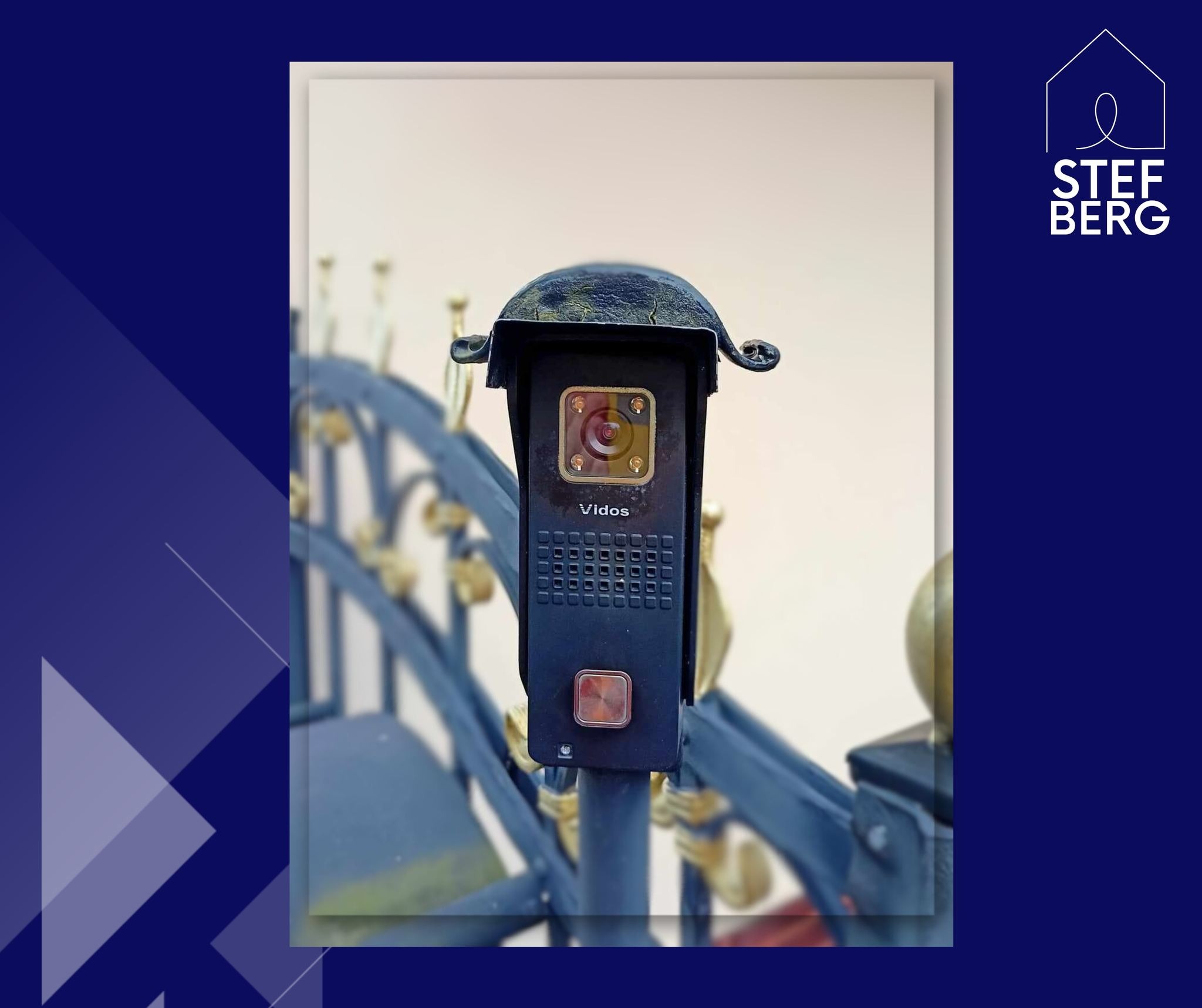 Czarny panel wejściowy z kamerą i przyciskiem dzwonka, model Vidos, zamontowany na metalowym słupku bramy z ozdobnymi elementami.