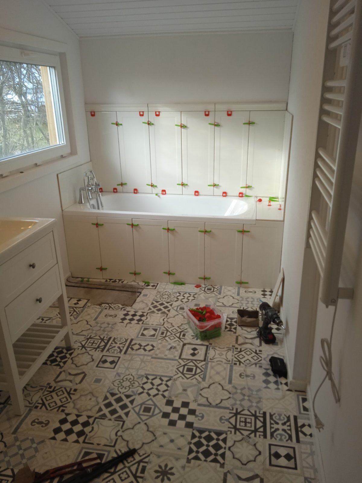 Biała wanna obudowana płytkami, z zamontowanymi klipsami poziomującymi, widok na wzorzystą podłogę z ułożonymi płytkami, pojemnik z czerwonymi klinami poziomującymi, wiertarka akumulatorowa.