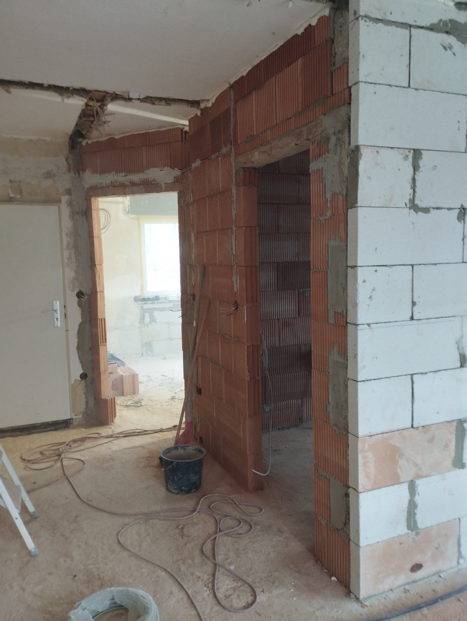 Wnętrze w trakcie przebudowy: widoczne ściany z czerwonej cegły i białych bloczków, fragment odsłoniętego sufitu, wiadro i kable na podłodze, w tle widok na kolejne pomieszczenie.