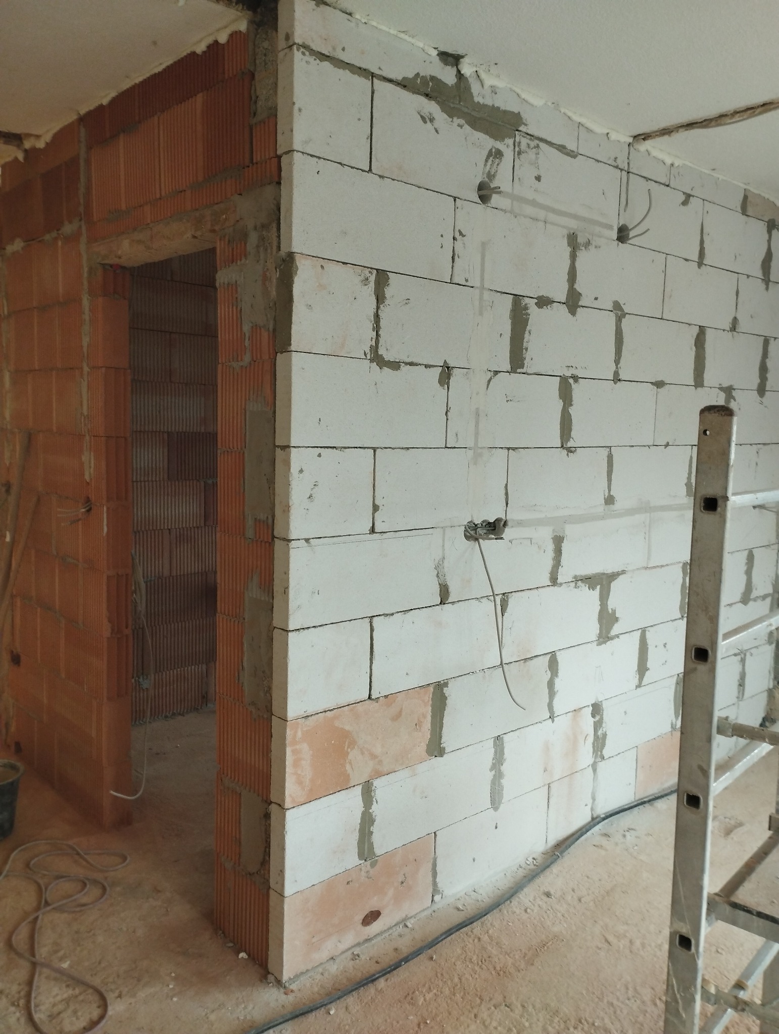 Murowana ściana z pustaków ceramicznych i betonu komórkowego w trakcie budowy, widoczne przewody elektryczne, fragment drabiny aluminiowej i niedokończone nadproże drzwiowe.