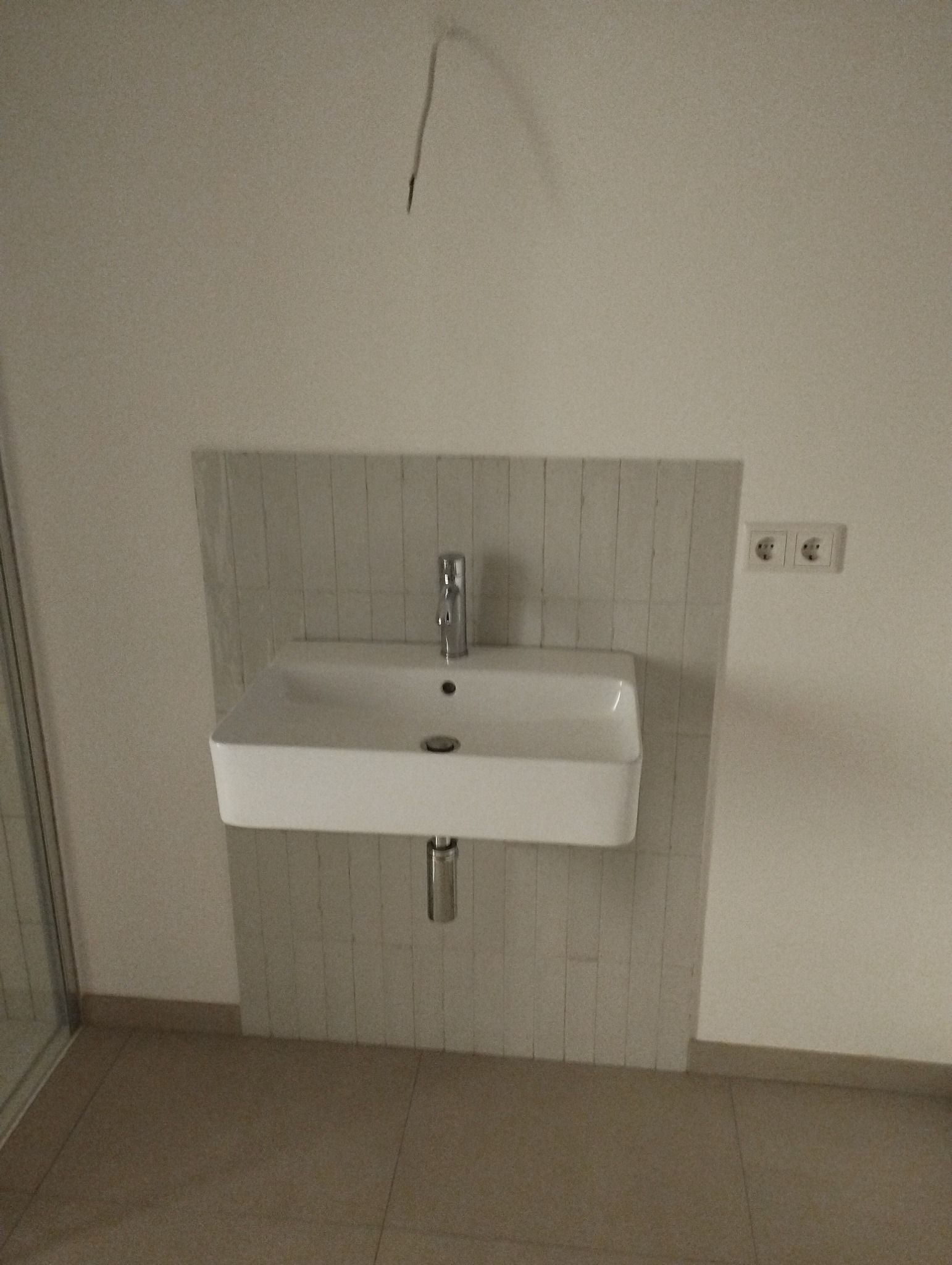 Nowoczesna, prostokątna umywalka zamontowana na ścianie wyłożonej pionowymi, jasnymi płytkami, z widocznym odpływem i baterią, w łazience w trakcie wykańczania, z wystającym kablem ze ściany.