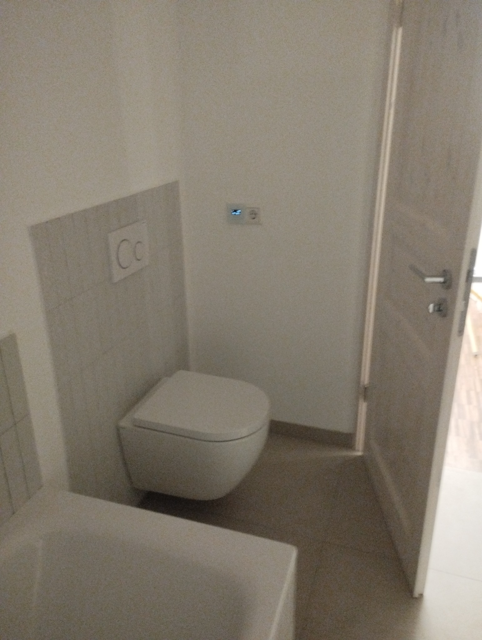 Nowoczesna, podwieszana toaleta w minimalistycznej łazience z beżowymi płytkami podłogowymi, widoczny fragment wanny oraz białe drzwi z metalową klamką.