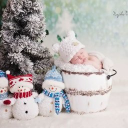 NAJKA SPÓŁKA Z OGRANICZONĄ ODPOWIEDZIALNOŚCIĄ - Śpiący noworodek w białym, futrzanym wiaderku obok pluszowych bałwanków i ośnieżonego drzewka, zimowa sceneria, delikatne pastelowe tło.