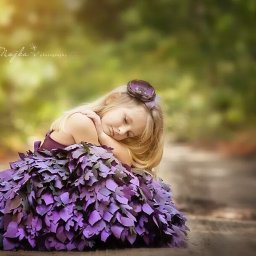 NAJKA SPÓŁKA Z OGRANICZONĄ ODPOWIEDZIALNOŚCIĄ - Śpiąca blond dziewczynka w fioletowej sukience z liści, leżąca na leśnej ścieżce, z fioletową ozdobą we włosach, w miękkim, rozproszonym świetle.