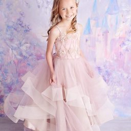 NAJKA SPÓŁKA Z OGRANICZONĄ ODPOWIEDZIALNOŚCIĄ - Uśmiechnięta dziewczynka w tiulowej, różowej sukience z aplikacjami w kształcie róż i koroną na głowie, pozuje na tle malowanego zamku.