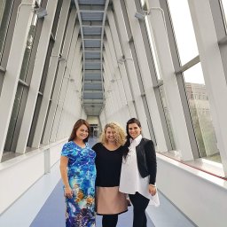 NAJKA SPÓŁKA Z OGRANICZONĄ ODPOWIEDZIALNOŚCIĄ - Trzy uśmiechnięte kobiety stojące w nowoczesnym korytarzu z ukośnymi oknami, kobieta po lewej w długiej sukience w morskie wzory, środkowa w czarnej bluzce i beżowej spódnicy, a po prawej w białej...