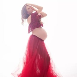 NAJKA SPÓŁKA Z OGRANICZONĄ ODPOWIEDZIALNOŚCIĄ - Kobieta w ciąży, w czerwonej sukni, pozuje na jasnym tle. Ujęcie w pełnej sylwetce, z delikatnym uśmiechem i rękami uniesionymi nad głową.