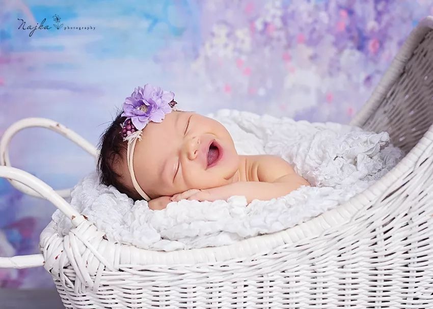 Śpiące niemowlę z otwartymi ustami, ubrane w opaskę z fioletowym kwiatem, leży w białym wiklinowym koszu na tle rozmytego, fioletowo-niebieskiego tła.