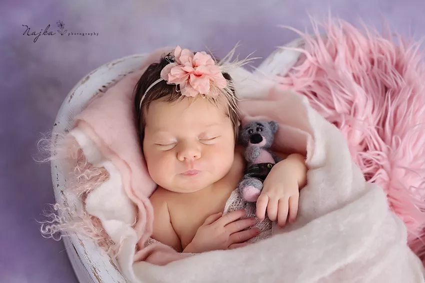 Śpiąca noworodka z różową opaską na głowie, otulona miękkim kocykiem w drewnianej wanience, przytulająca małego pluszowego misia.