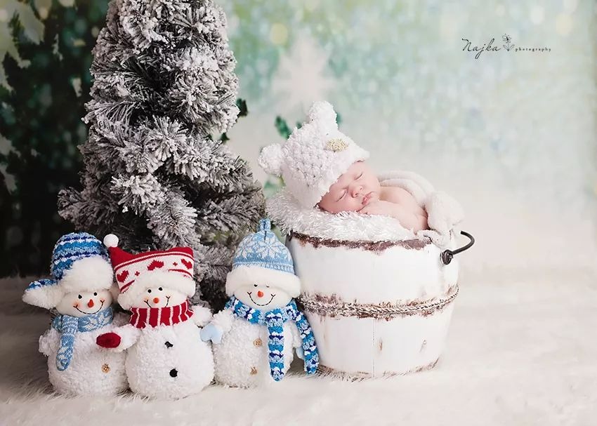 Śpiący noworodek w białym, futrzanym wiaderku obok pluszowych bałwanków i ośnieżonego drzewka, zimowa sceneria, delikatne pastelowe tło.