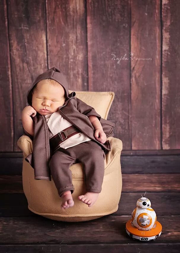 Śpiący noworodek w kostiumie Jedi z filmu Gwiezdne Wojny, siedzący w beżowym fotelu, obok figurka BB-8 na tle drewnianej ściany.