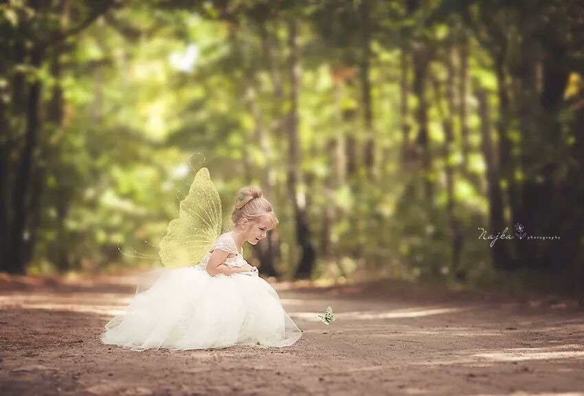 Dziewczynka w białej sukience i ze skrzydłami motyla siedzi na leśnej ścieżce, obserwując motyla.