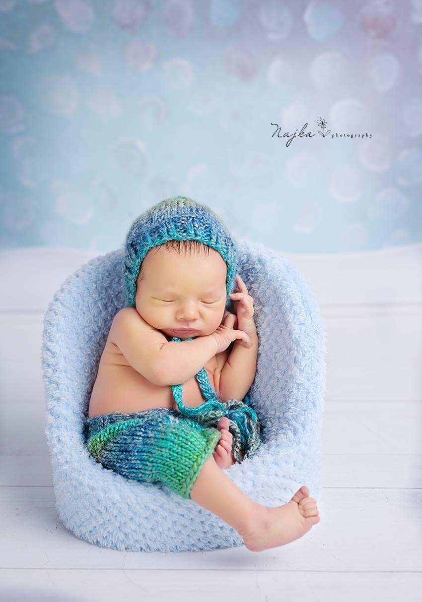 Śpiący noworodek w niebiesko-zielonym, dzierganym ubranku, ułożony w miękkim, błękitnym foteliku na tle rozmytego, błękitnego tła z efektem bokeh.