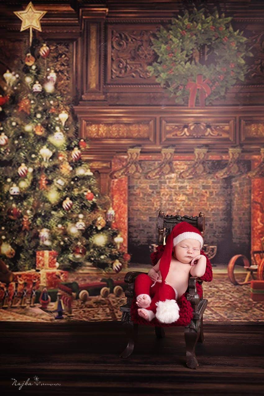 Śpiący noworodek w czerwonym stroju Mikołaja, siedzący w krześle na tle świątecznym z choinką i kominkiem, wykonane w studio fotograficznym w Warszawie.