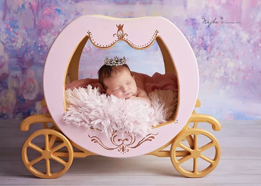 Śpiąca noworodka w koronie, ułożona w różowo-złotej, dekoracyjnej karocy na tle pastelowej, kwiatowej ściany.