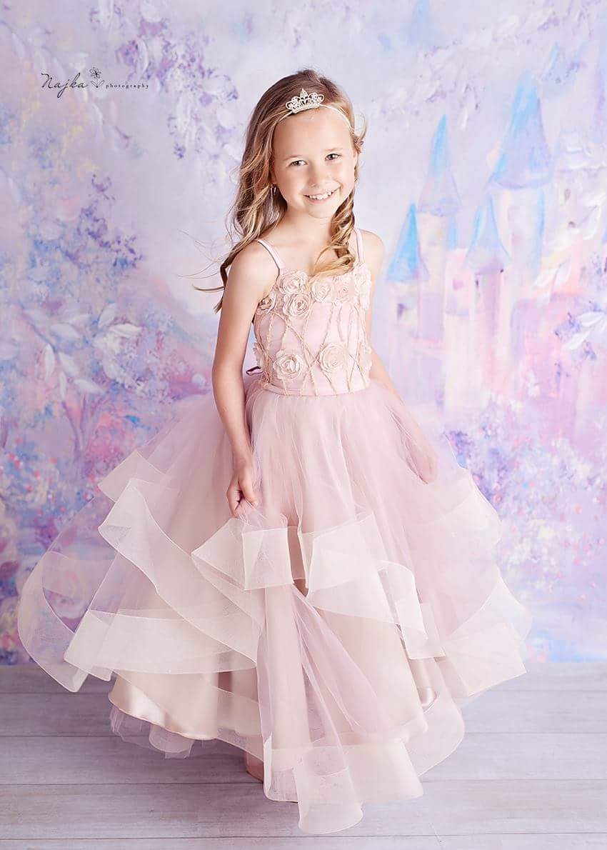 Uśmiechnięta dziewczynka w tiulowej, różowej sukience z aplikacjami w kształcie róż i koroną na głowie, pozuje na tle malowanego zamku.
