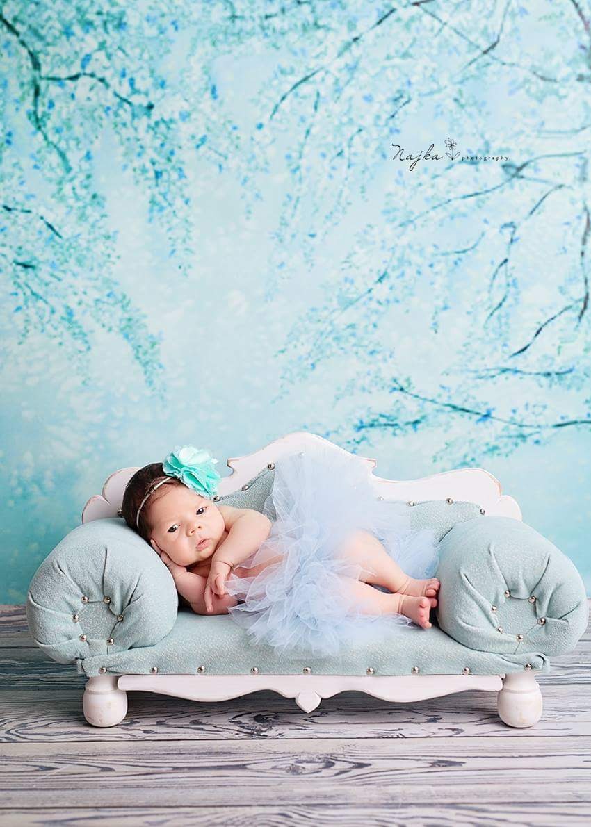 Noworodek w błękitnej tiulowej spódniczce i opasce z kwiatem, leżący na małej, pastelowej kanapie na tle błękitnego, kwiecistego tła studyjnego.