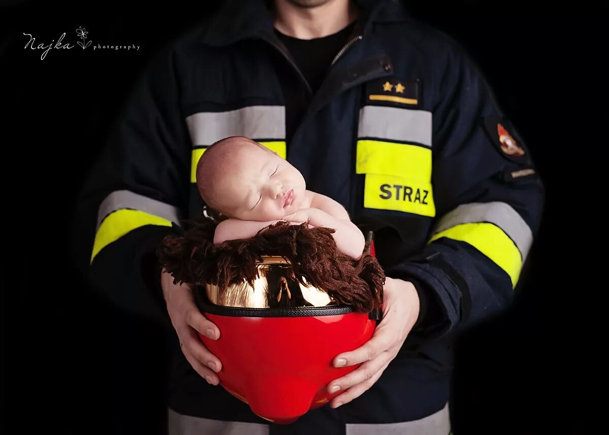 Śpiący noworodek ułożony w czerwonym hełmie strażackim, trzymanym przez mężczyznę w mundurze strażackim na czarnym tle.