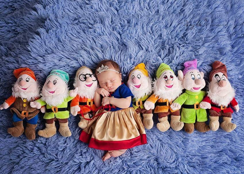 Noworodek w stroju Królewny Śnieżki śpi otoczony pluszowymi krasnalami na niebieskim, puszystym tle.