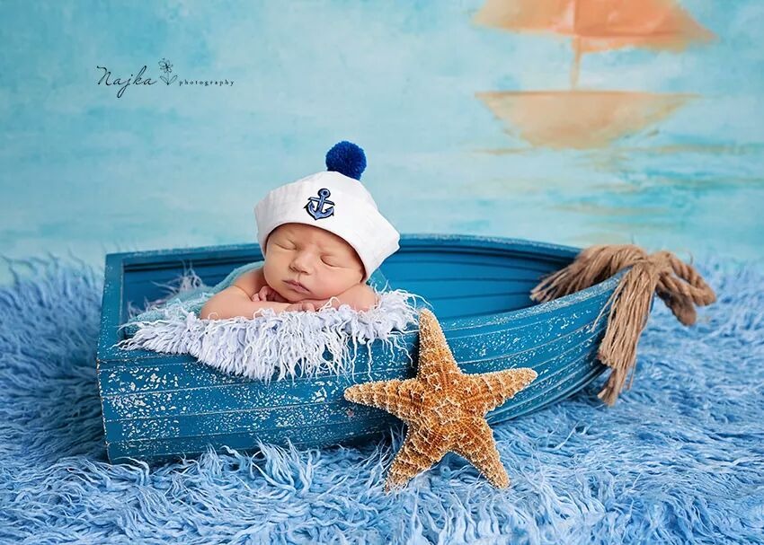 Śpiący noworodek w marynarskiej czapce z kotwicą, leżący w małej, błękitnej łódce na tle pomalowanego morza i żaglówki, obok leży rozgwiazda. Stylizacja morska.