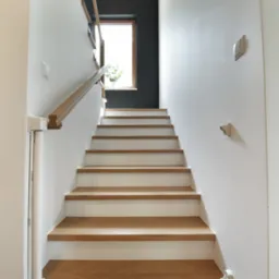 Nowoczesne schody z drewnianymi stopniami i białymi podstopnicami, drewniana poręcz, widok z dołu na górę, ściany białe i antracytowe, okno z rośliną w tle.