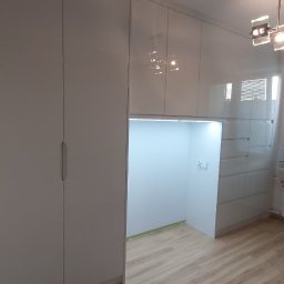ORGANMISTRZOWIE SPÓŁKA Z OGRANICZONĄ ODPOWIEDZIALNOŚCIĄ - Biała szafa wnękowa z połyskiem, wbudowana w ścianę, z oświetloną wnęką i drewnianą podłogą, nowoczesne oświetlenie sufitowe.