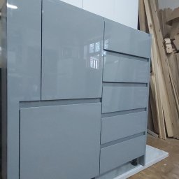 ORGANMISTRZOWIE SPÓŁKA Z OGRANICZONĄ ODPOWIEDZIALNOŚCIĄ - Szara, lakierowana komoda z szufladami i szafkami, stojąca na białym cokole, widoczna w warsztacie stolarskim z drewnem w tle.