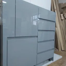 Szara, lakierowana komoda z szufladami i szafkami, stojąca na białym cokole, widoczna w warsztacie stolarskim z drewnem w tle.