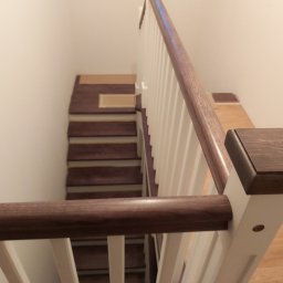 ORGANMISTRZOWIE SPÓŁKA Z OGRANICZONĄ ODPOWIEDZIALNOŚCIĄ - Widok z góry na drewniane schody z białymi balustradami i mahoniowymi poręczami, częściowo zabezpieczone kartonem podczas prac wykończeniowych.