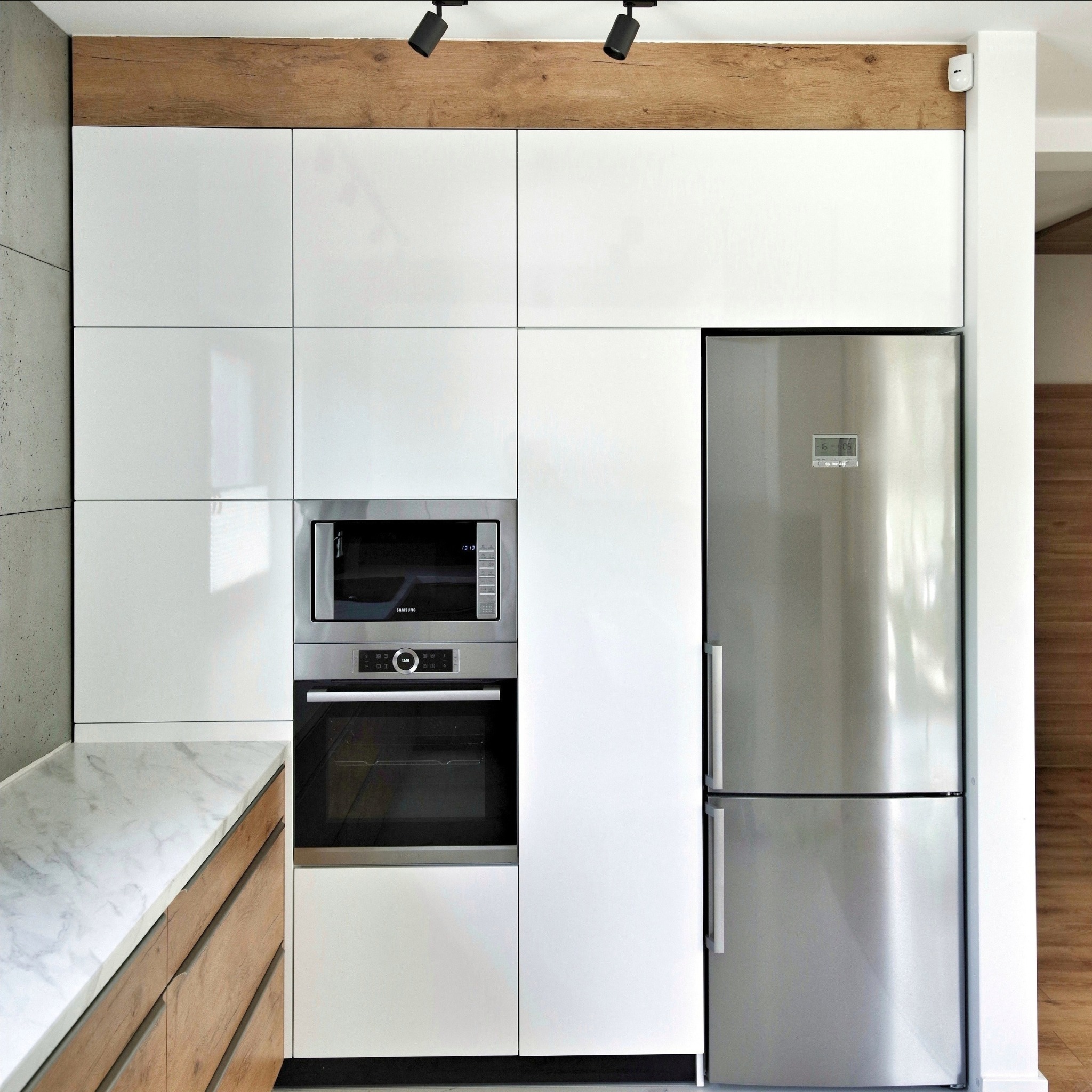 Nowoczesna zabudowa kuchenna z białymi, lakierowanymi frontami, wbudowanym piekarnikiem i mikrofalówką, oraz lodówką side-by-side ze stali nierdzewnej, uzupełniona blatem z imitacją marmuru...