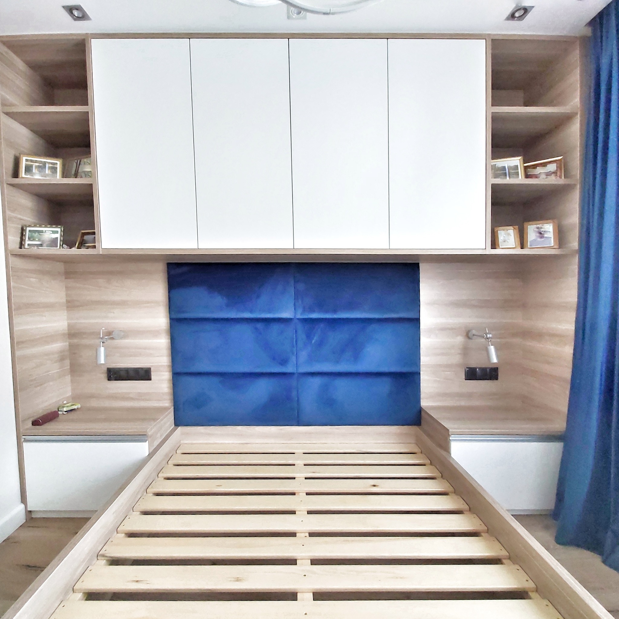 Drewniana rama łóżka z niebieskim panelem wezgłowia, zabudowana szafkami nocnymi z lampkami i szafkami wiszącymi z półkami na zdjęcia.