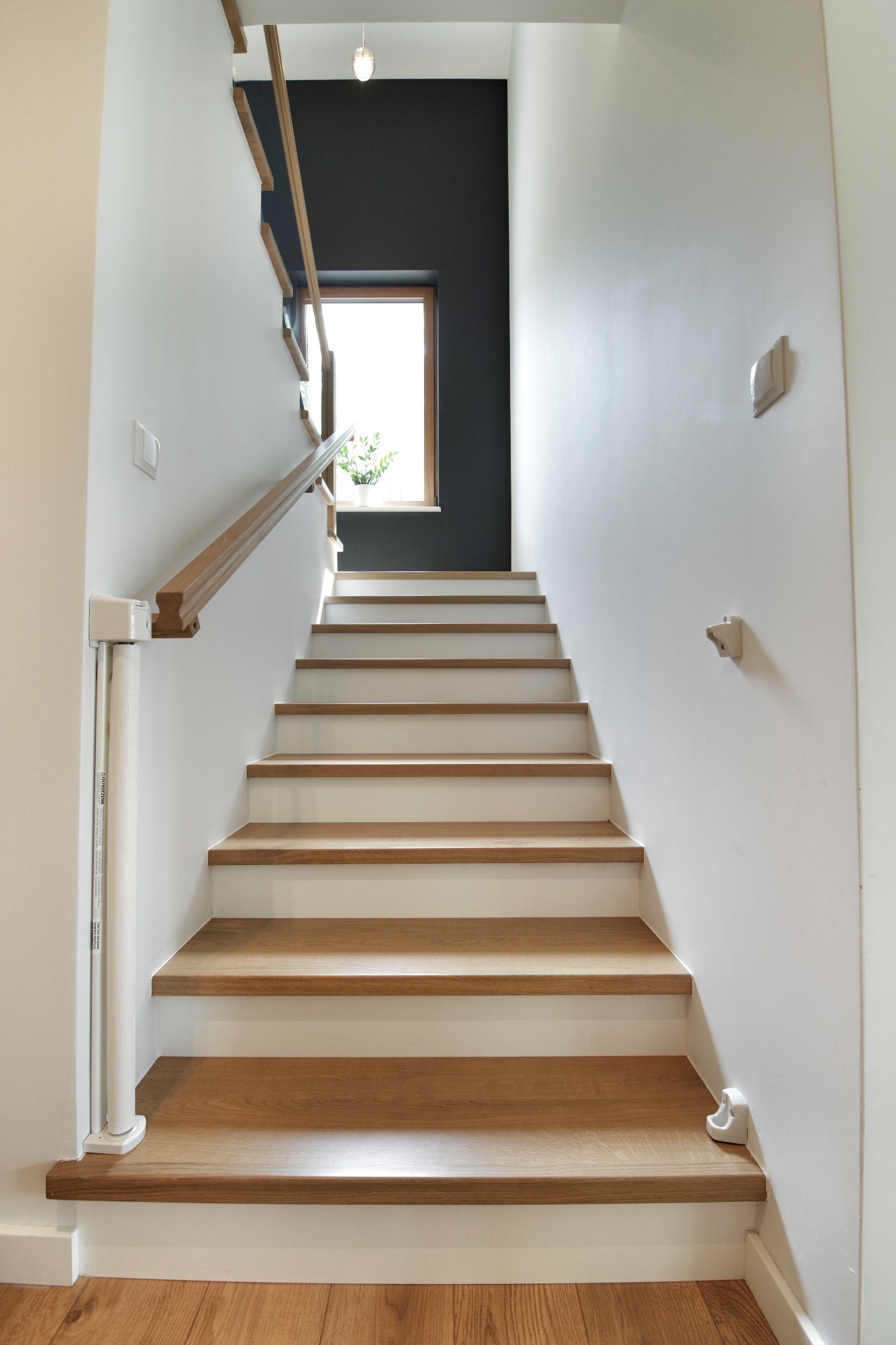 Nowoczesne schody z drewnianymi stopniami i białymi podstopnicami, drewniana poręcz, widok z dołu na górę, ściany białe i antracytowe, okno z rośliną w tle.