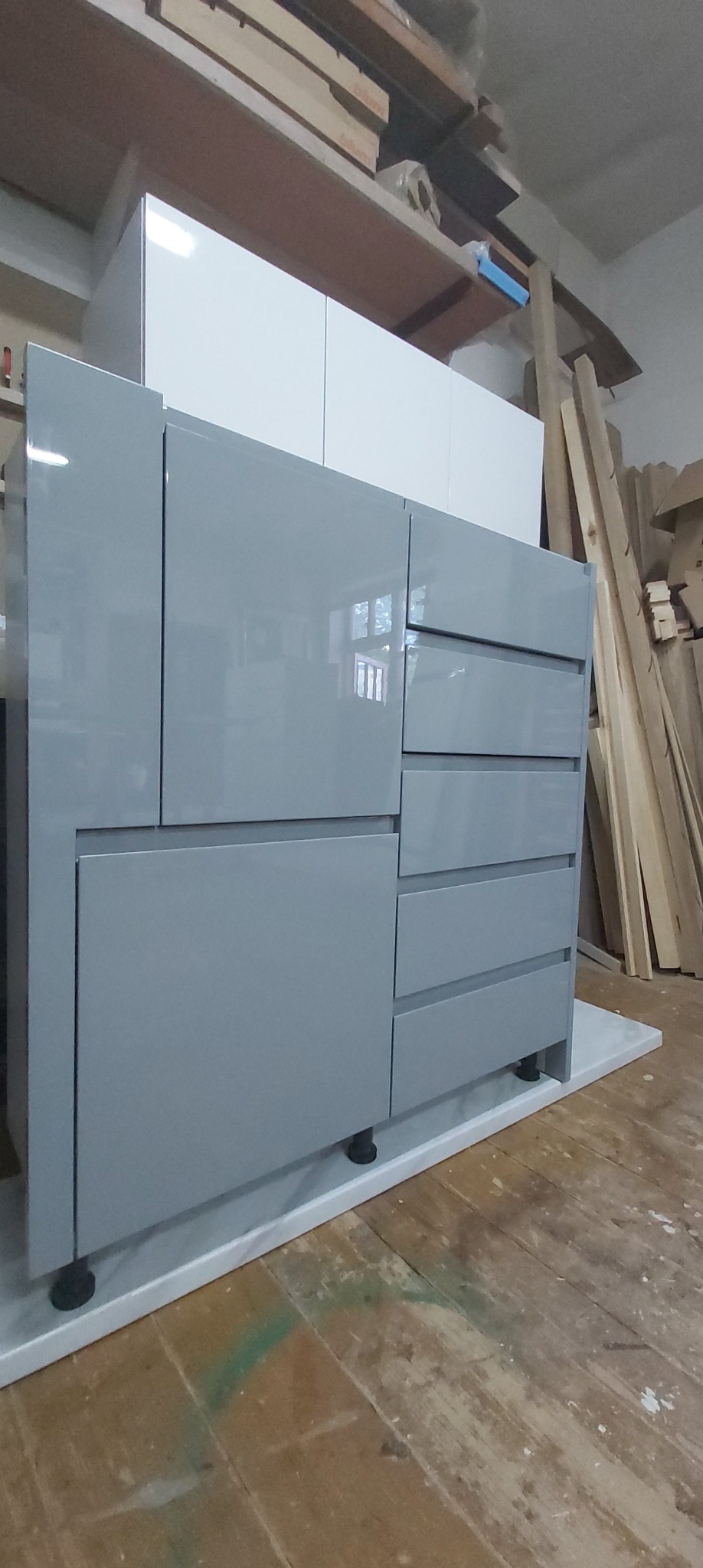 Szara, lakierowana komoda z szufladami i szafkami, stojąca na białym cokole, widoczna w warsztacie stolarskim z drewnem w tle.