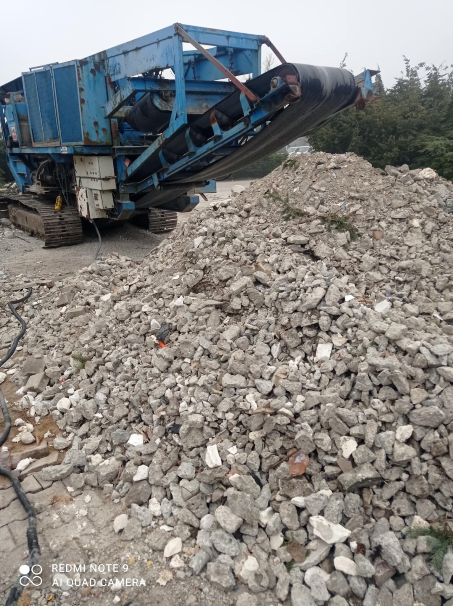 Niebieski kruszarkowy przesiewacz gąsienicowy przerabia gruz betonowy na placu budowy. W tle drzewa.