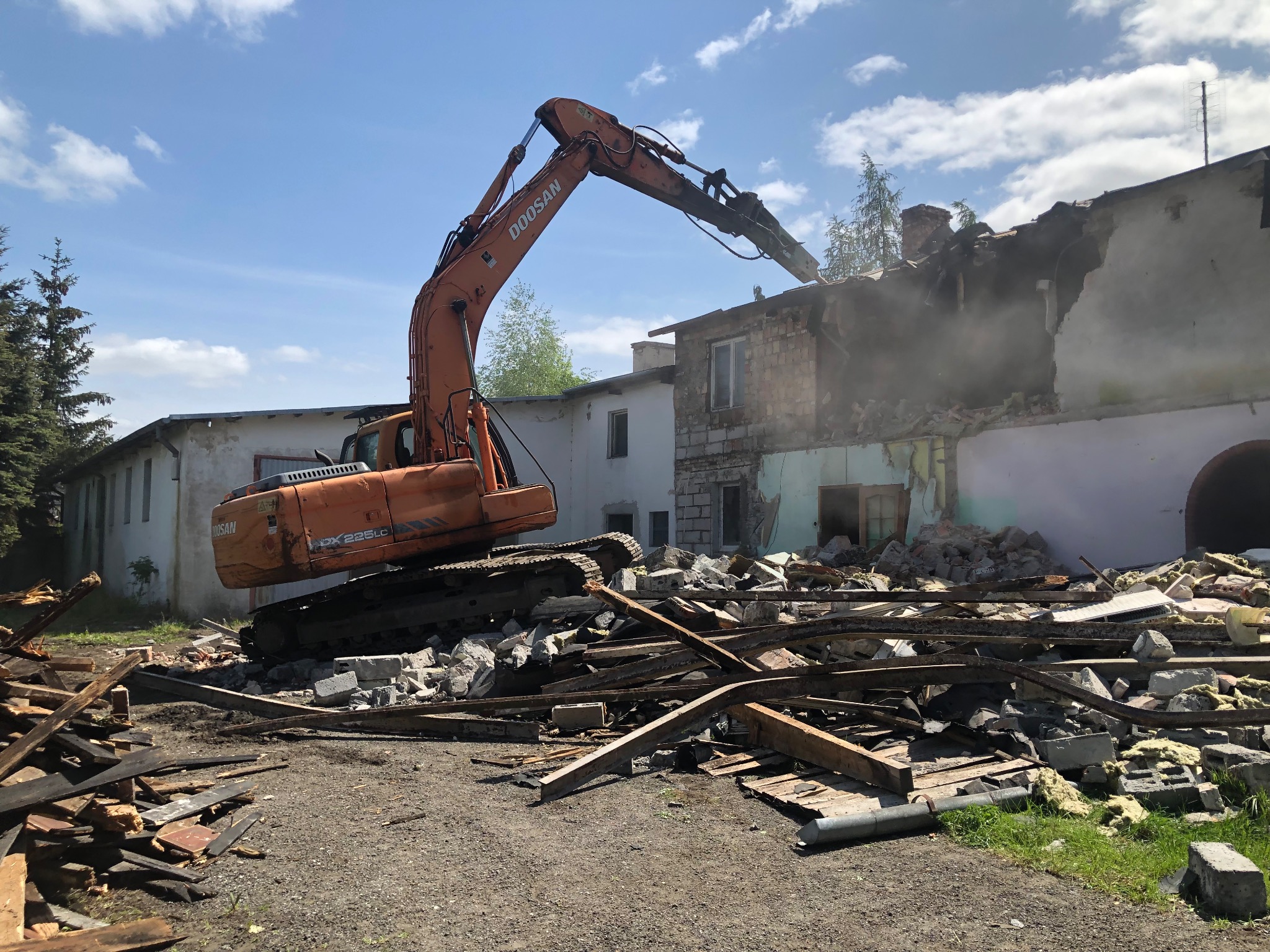 Pomarańczowa koparka Doosan w trakcie rozbiórki budynku; gruz i drewniane elementy na pierwszym planie, budynek częściowo zburzony w tle.